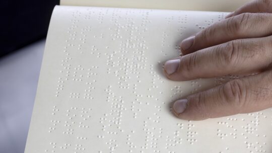 Bild eines Buches in Brailleschrift mit einer Hand darüber.